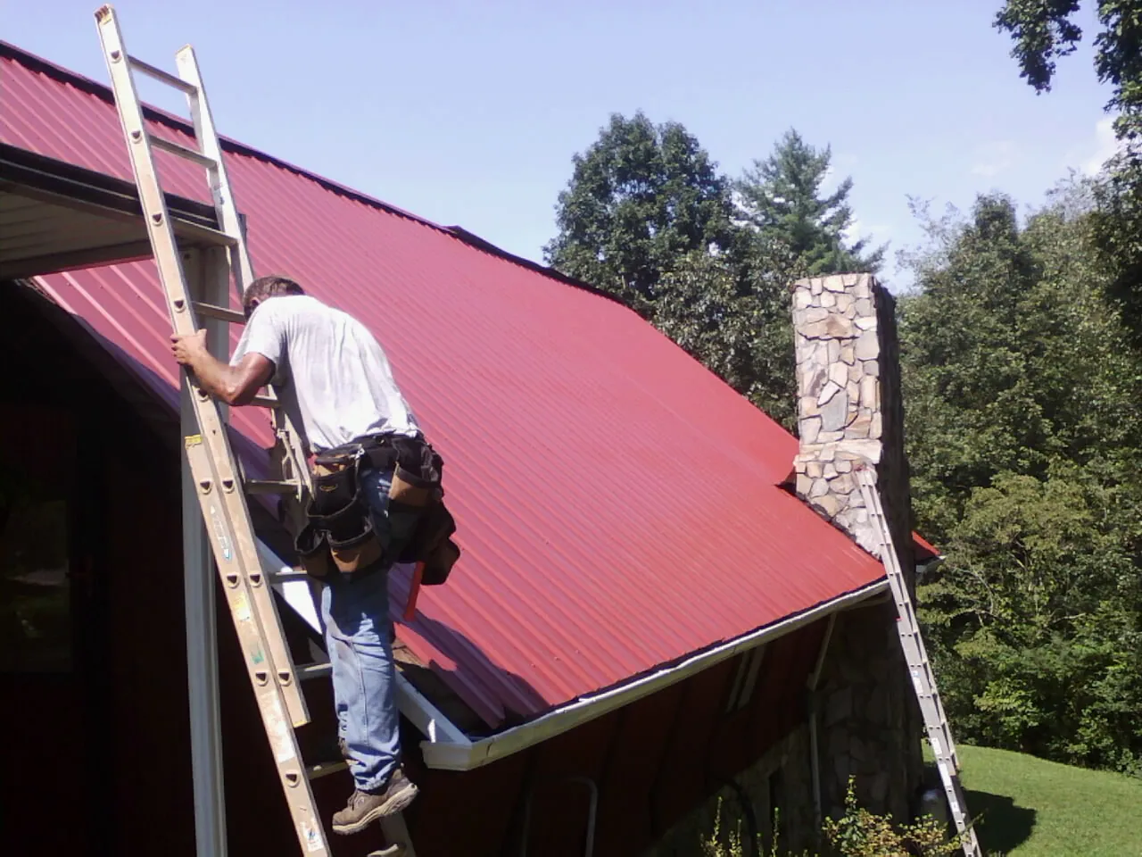 Roofer on a ladder beside a red metal roof and stone chimney — Soffit & Fascia Repair by Roofing Brothers in Mountain Lakes